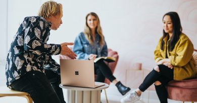 man in black and white floral long sleeve shirt sitting and showing something on a microsoft laptop to 2 women